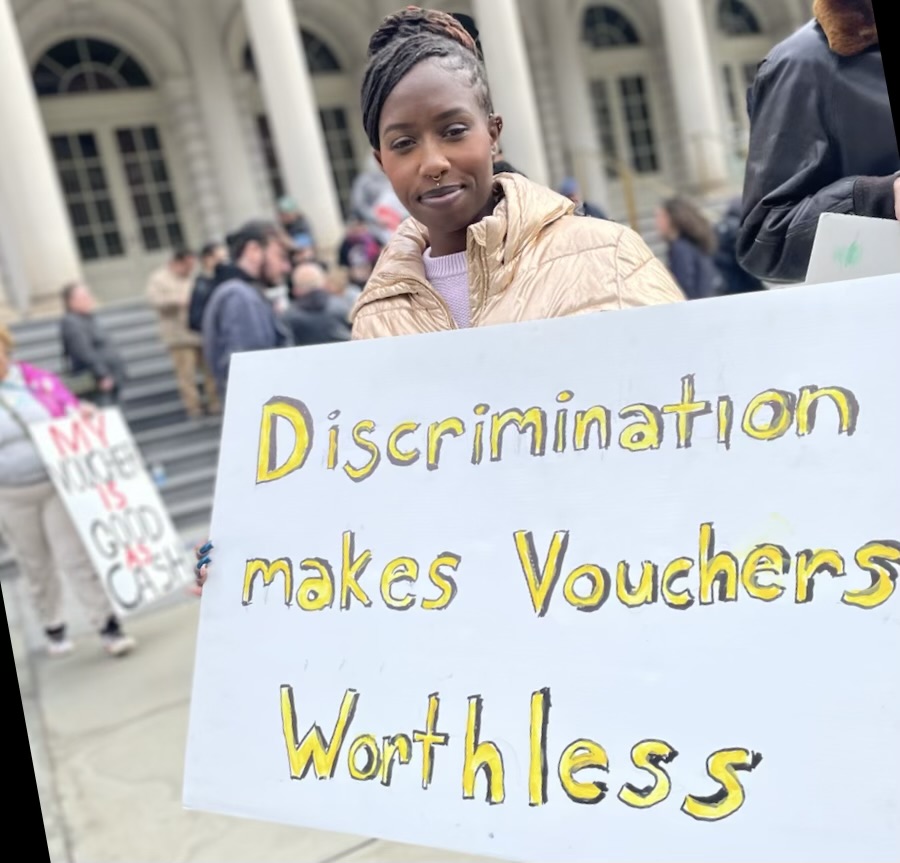 a young woman holding a handmade poster that says "Discrimination makes Vouchers Worthless" in front of city hall